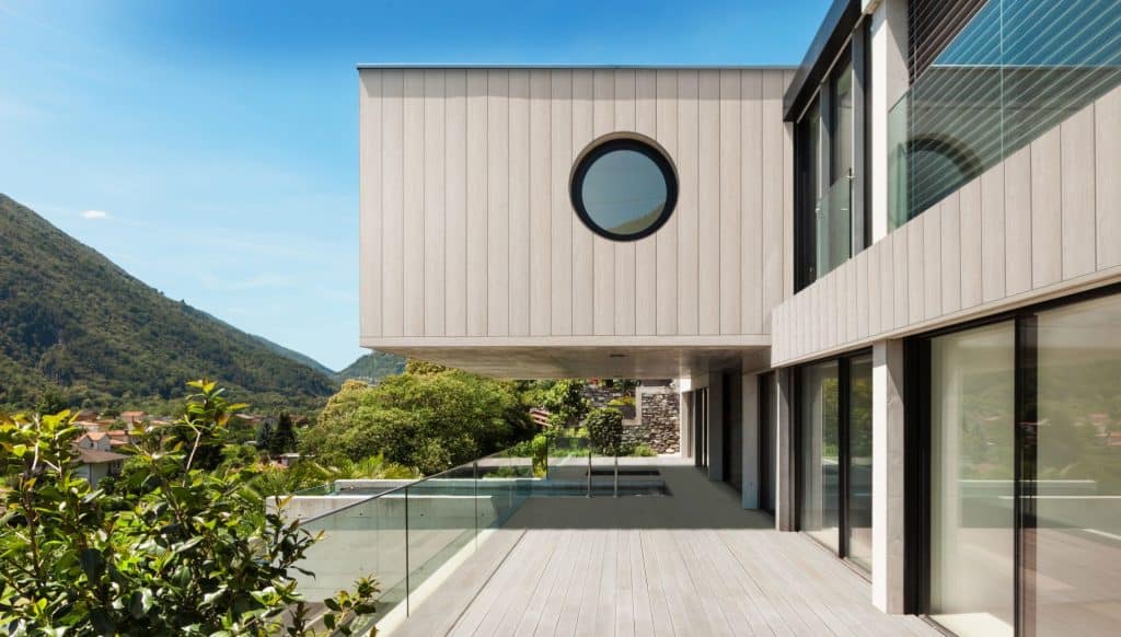 Modern home exterior with Millboard Cladding Envello Shadow Line+, circular window, and glass balcony overlooking a mountain view.